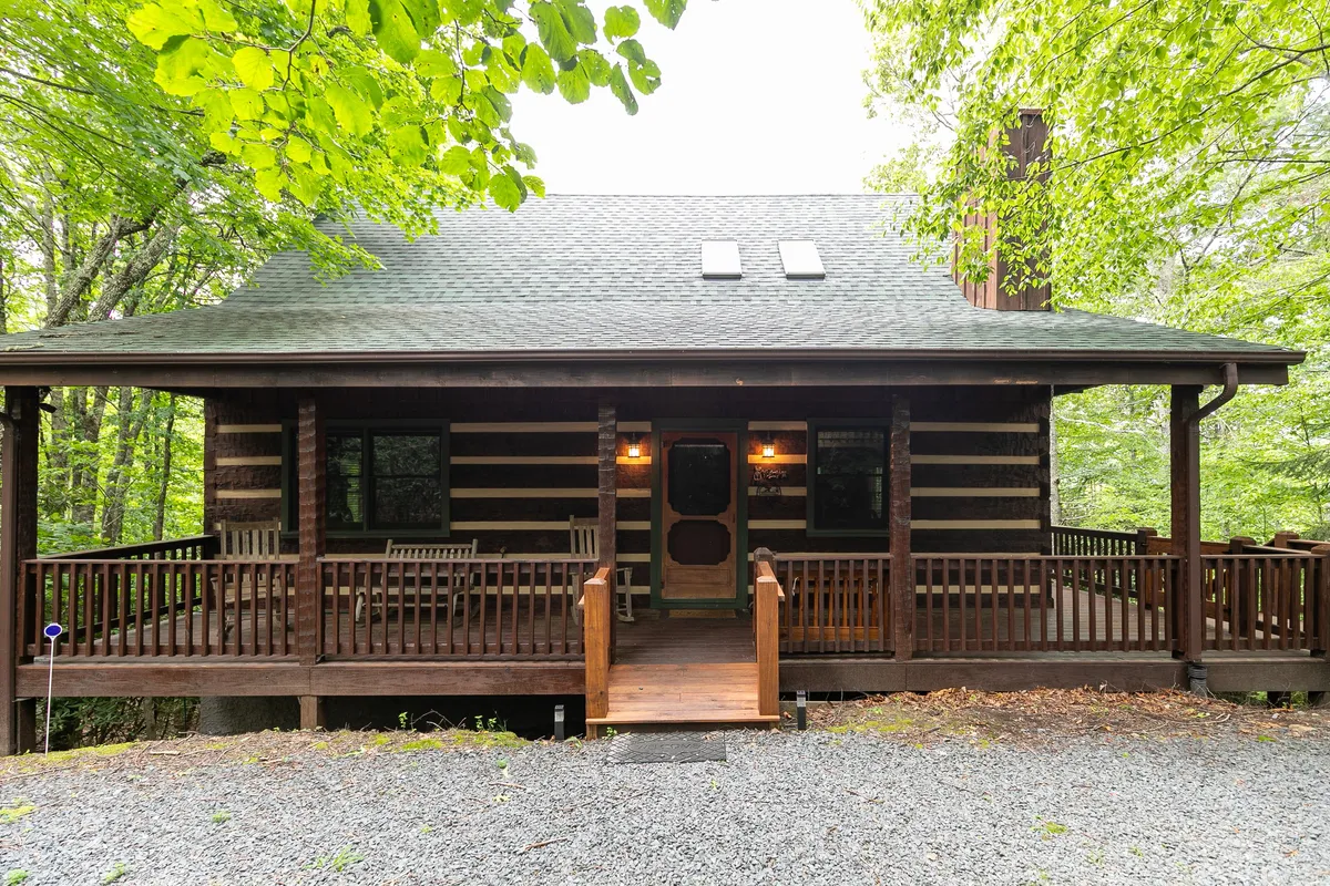 The image showcases a charming log cabin with a spacious wooden porch, surrounded by lush greenery. The porch offers sweeping sunset views and an inviting space for morning coffee or evening relaxation. The no-step entry and flat parking are subtly visible, emphasizing easy accessibility. The scene feels tranquil and cozy, highlighting the cabin’s connection to nature and its suitability for gatherings or quiet retreats.