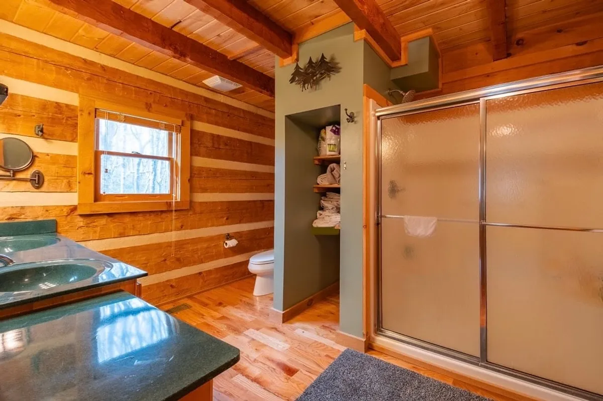 A spa-inspired bathroom featuring a double vanity with dual sinks, elegant wooden beams, a shower stall, and shelves stocked with plush towels. Natural light streams in through the window, highlighting the room’s spaciousness and warmth.
