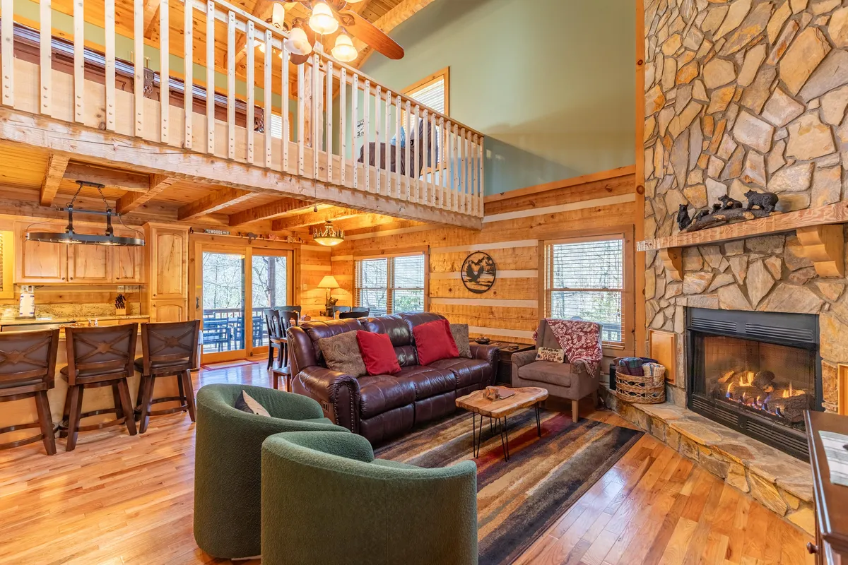 A beautiful great room with a dramatic two-story stone fireplace, leather sofa, exposed wooden beams, a balcony overlook, and large windows letting in natural light. The space is arranged for family gatherings and relaxation.