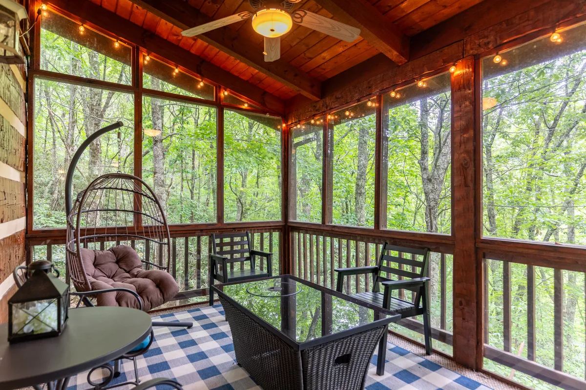 A cozy, glassed-in deck area features a hanging egg chair, a glass-top table, string lights, and wooden walls. The scene is serene and inviting, perfect for sipping coffee in the morning or sharing a quiet evening together while watching the sunset or stargazing.
