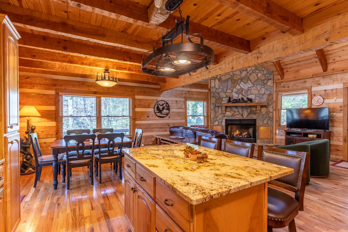 A wide-angle view of an open-concept living area featuring a granite kitchen island, stone fireplace, dining table, and rustic wooden beams. The setup highlights the seamless flow between kitchen, dining, and living spaces.