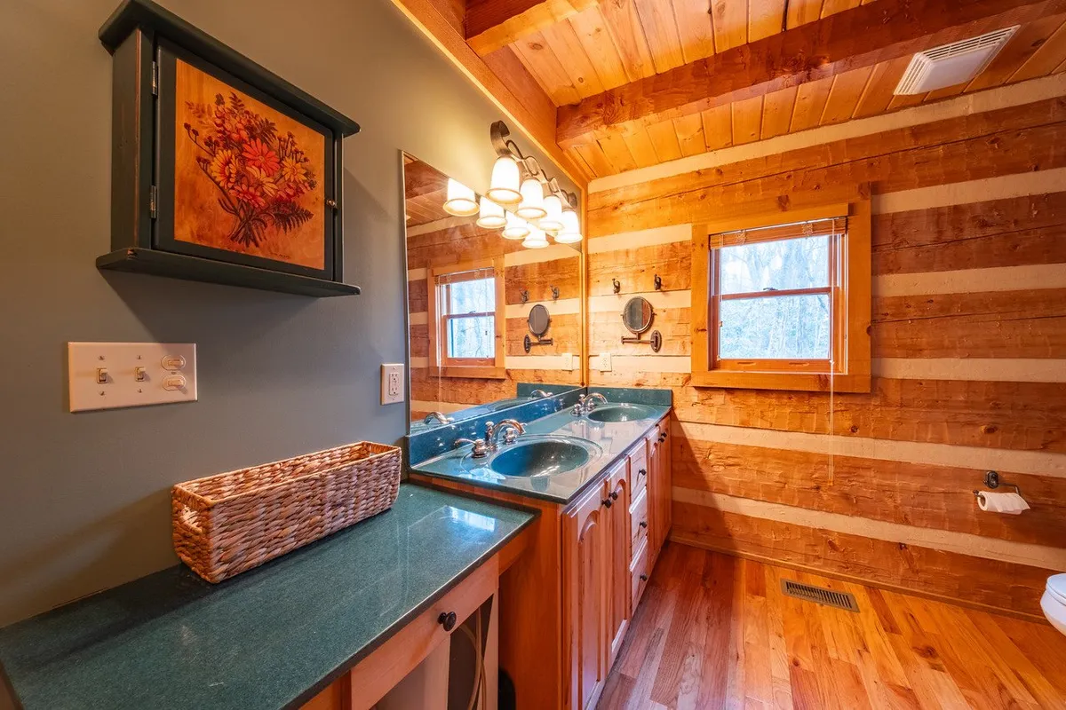 A cozy bathroom with a double sink vanity, wooden ceiling beams, large mirror, and framed artwork on the walls. The warm tones and morning light create a tranquil, spa-like feel ideal for self-care routines.