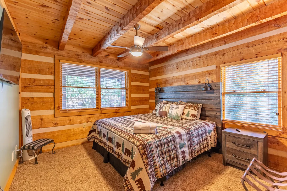 A cozy bedroom with patterned bedding, warm wooden walls and ceiling, and sunlight streaming in through multiple windows. The natural light highlights rustic textures and creates a peaceful, retreat-like atmosphere. The bed is perfectly made, inviting relaxation and comfort.