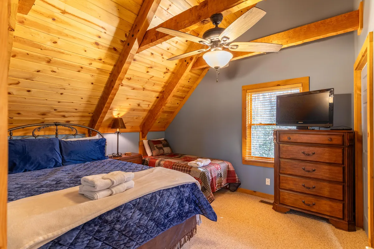 A bright and comfortable bedroom featuring a bed with blue bedding, a chest of drawers, wooden beams on the ceiling, and a window letting in soft, natural light. The room feels cozy and practical, offering both relaxation and ample storage.