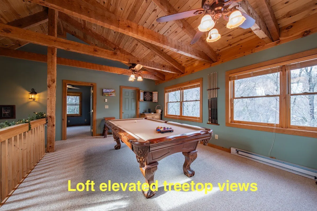 A stylish upstairs play loft featuring a full-size pool table set against large windows that offer treetop views. The space is accented by wooden ceiling beams, creating an airy, sunlit feel with plenty of space for social gatherings and relaxed recreation.