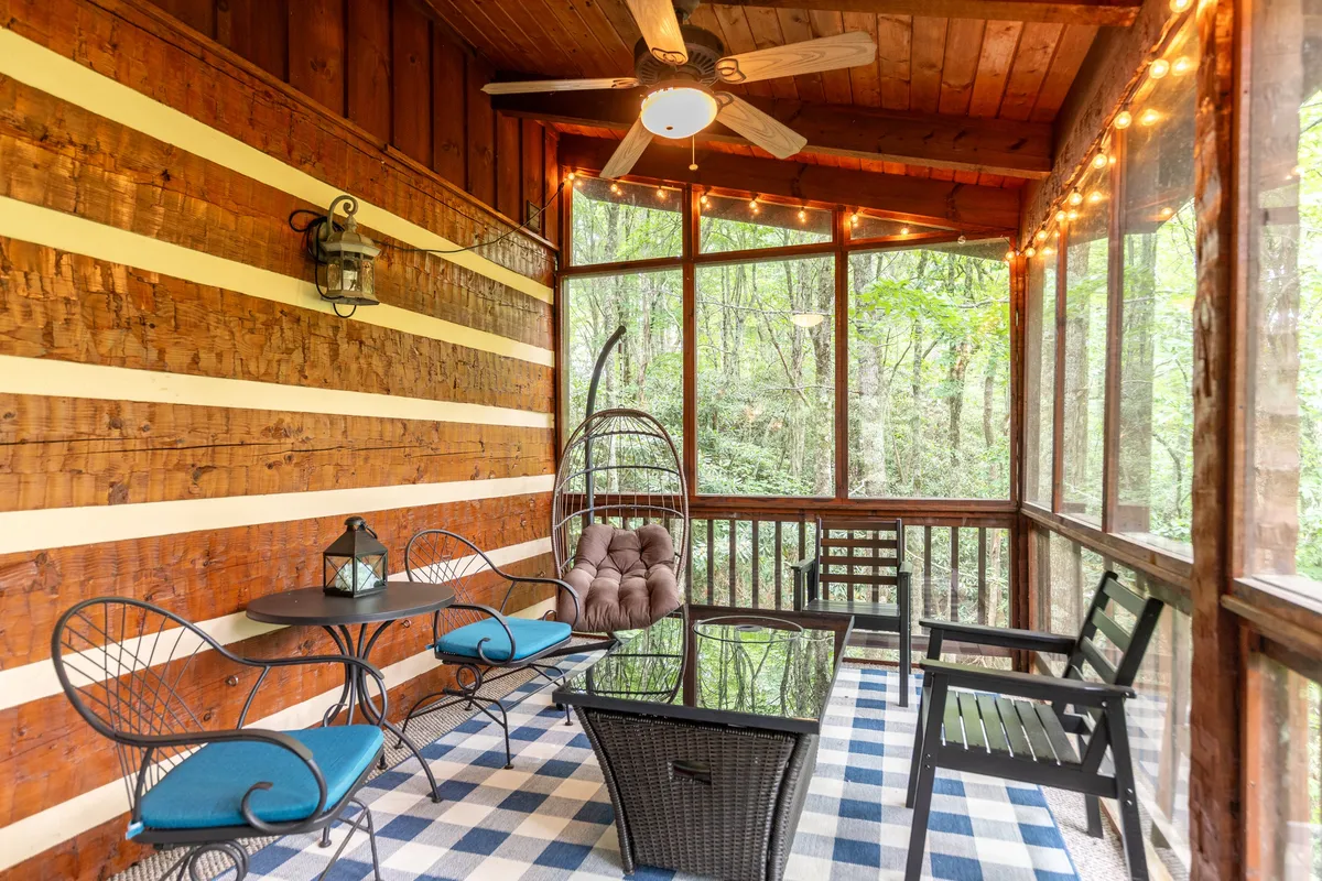 A cozy glassed-in porch featuring a signature egg swing chair, a small table with a lantern, plaid flooring, and wooden walls. The space is designed for relaxation and comfort, inviting guests to sway gently while enjoying a coffee, book, or conversation, protected from the elements.