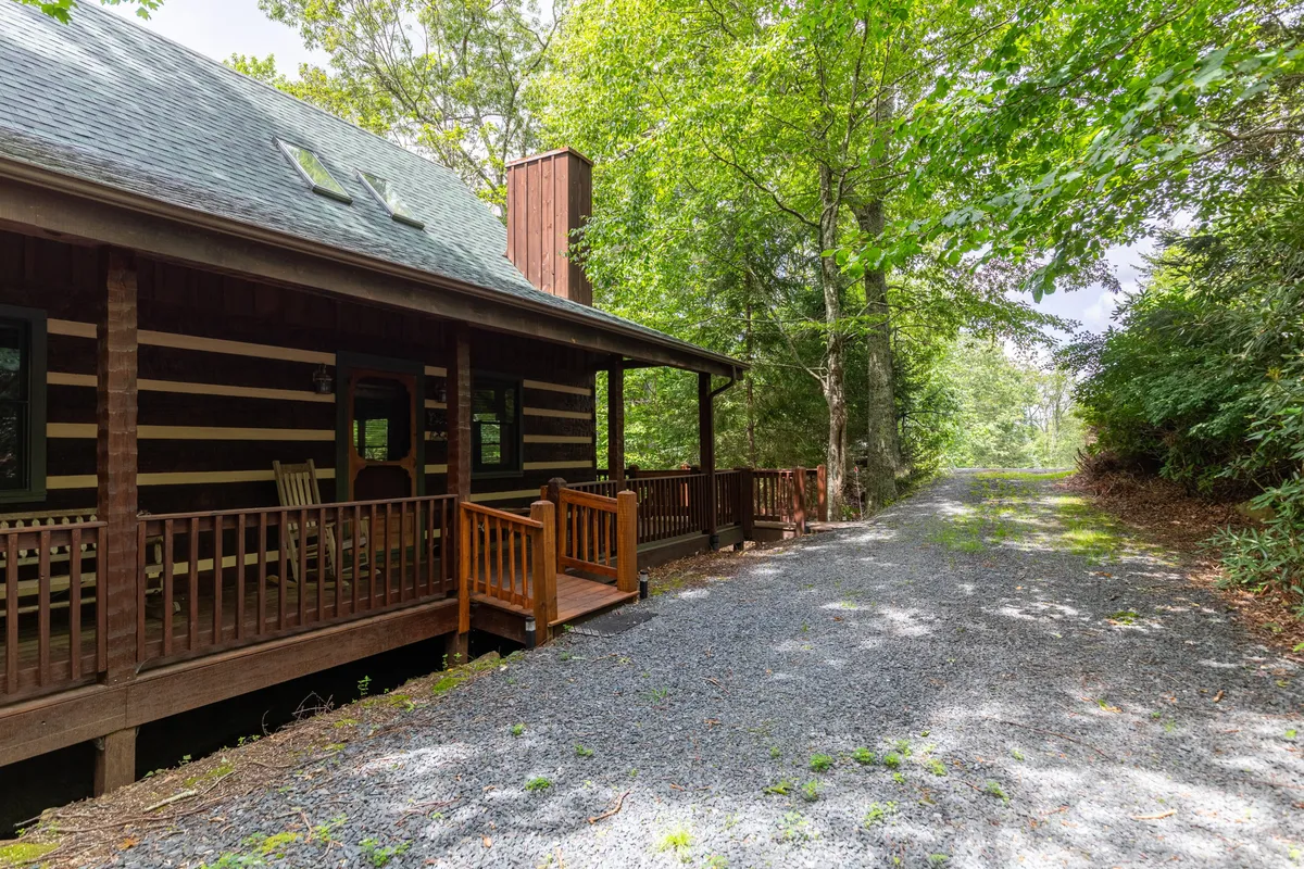 An exterior view of the log cabin, featuring a welcoming porch, gravel pathway, and a backdrop of mature trees. The scene conveys a sense of rustic serenity, with ample space for casual walks and outdoor enjoyment for both people and pets.