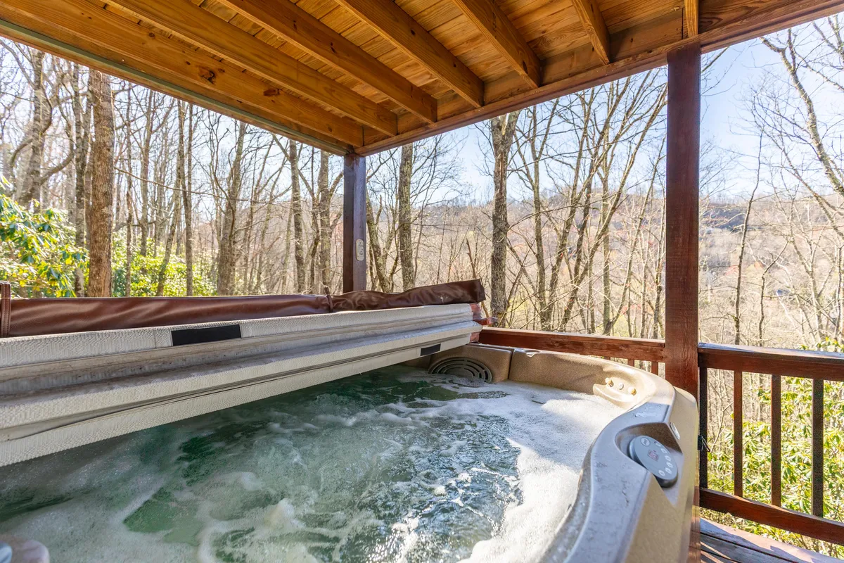 A bubbling hot tub sits on the covered deck, overlooking lush hills and surrounded by trees. The scene is serene yet social, suggesting relaxation after a day of adventure, with enough space for several people to enjoy the view together.