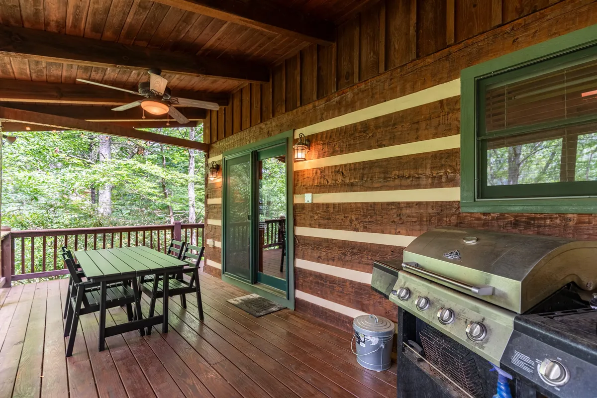 An inviting wooden deck outfitted with a spacious outdoor dining table and a grill, surrounded by lush greenery. The deck features a wooden ceiling, providing shelter while allowing for year-round use. The scene conveys a relaxed, cozy atmosphere perfect for alfresco dining and social gatherings.