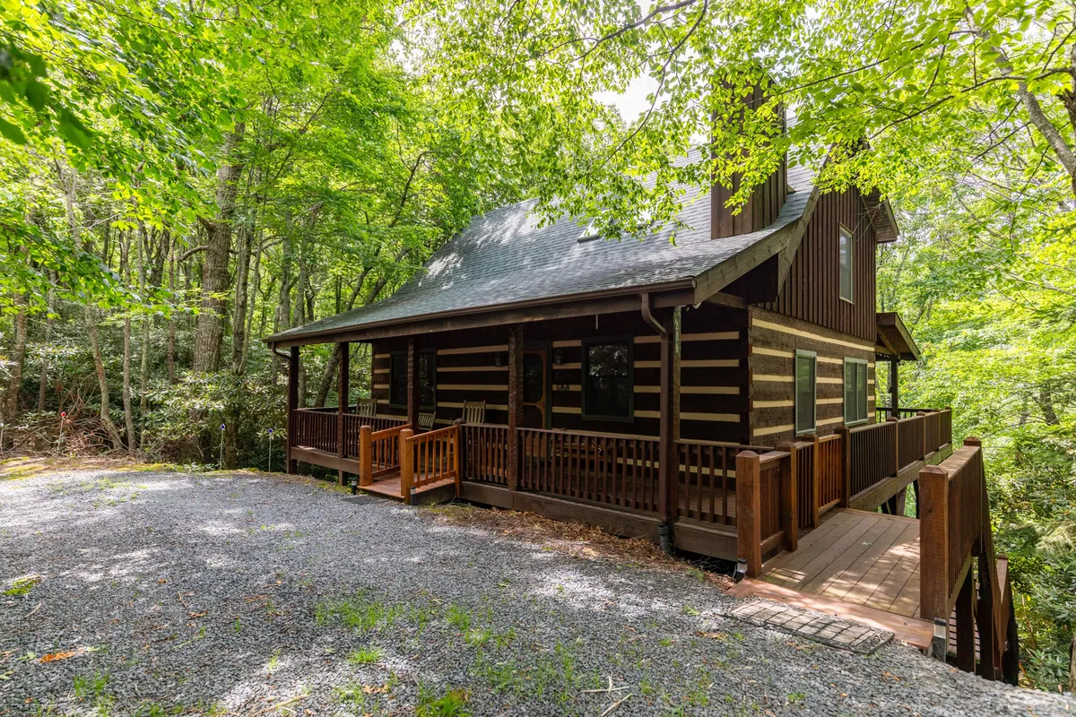A log cabin structure with a welcoming wooden porch, surrounded by tall trees. The gravel driveway in front offers ample space for parking, while the porch is an inviting place for gathering. The image captures the cabin's connection to the outdoors, emphasizing a communal, social atmosphere.
