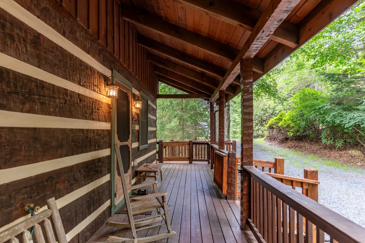 A cozy, covered front porch with classic wooden rocking chairs, log cabin structure, lamps on the walls, and lush greenery surrounding the entrance. The flat pathway and spacious entry area are clearly visible, emphasizing easy access.