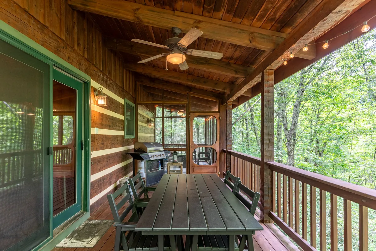 A spacious covered deck with a wooden dining table, outdoor grill, string lights, and a railing overlooking the trees. The setup is ideal for outdoor family meals, barbecues, and relaxed evenings together.