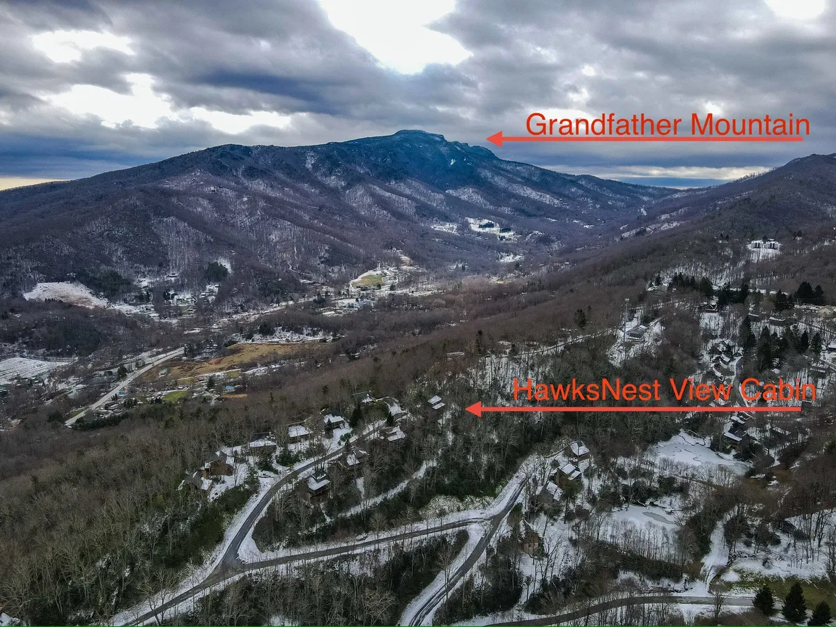 A wide, scenic view featuring HawksNest View Cabin nestled among trees, Grandfather Mountain in the background, and a snow-covered landscape with winding roads. The image suggests both winter adventure and tranquil relaxation, showing the cabin as a cozy base for multi-generational family activities.